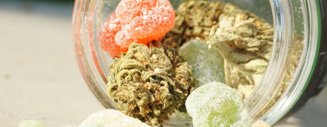 Glass jar spilling out colorful CBD gummies and cannabis buds on a table in natural sunlight.