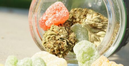 Glass jar spilling out colorful CBD gummies and cannabis buds on a table in natural sunlight.
